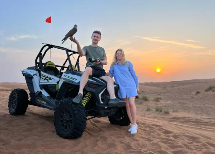 Two guests in helmets drive a dune buggy across Dubai’s red dunes at sunset, sand arcing behind them with a 4×4 on a nearby ridge.