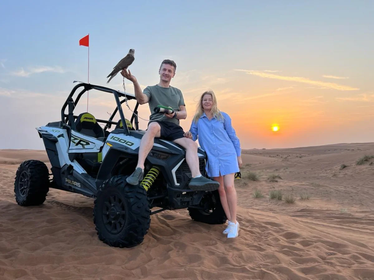 Two guests in helmets drive a dune buggy across Dubai’s red dunes at sunset, sand arcing behind them with a 4×4 on a nearby ridge.